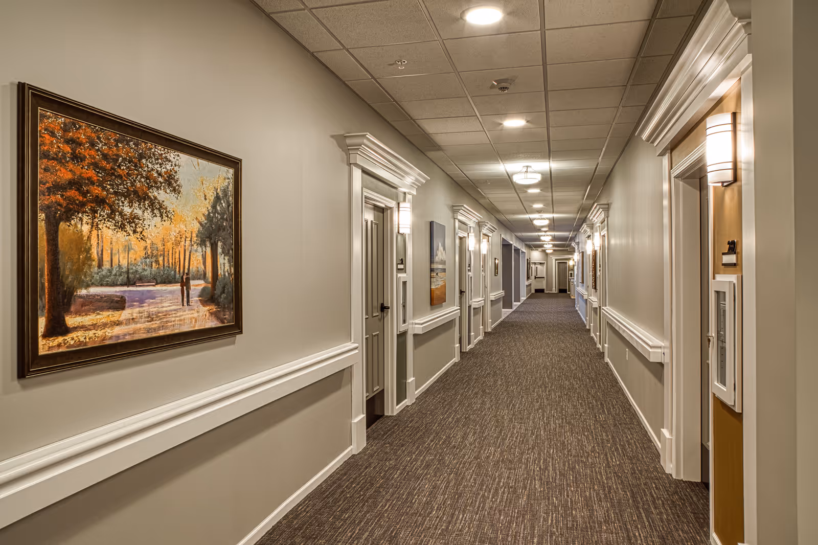 Long carpeted hallway in a senior living facility lined with doors, wall sconces, and framed artwork.