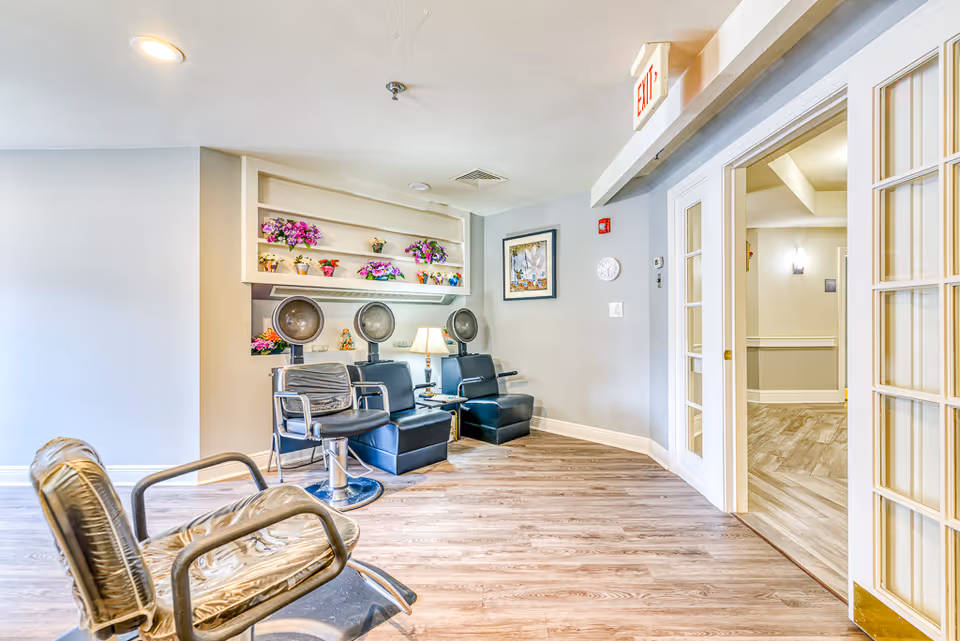 Salon area in a senior living facility featuring hair-dryer stations, styling chairs, shelves with flowers, and an open doorway.
