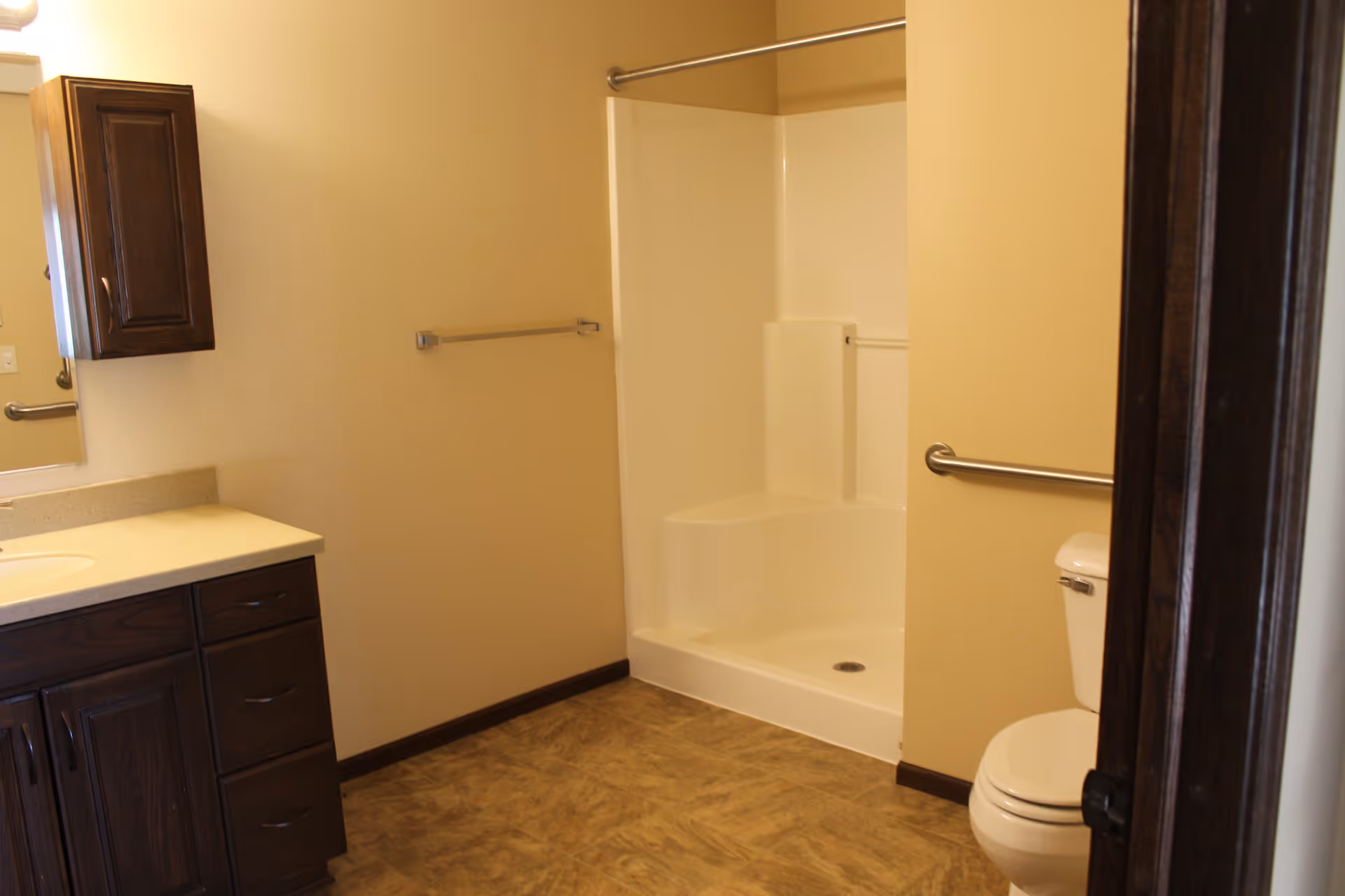 A bathroom with beige walls and flooring, featuring a white shower with a built-in seat and grab bars, a white toilet with a grab bar beside it, and a dark wood vanity with a beige countertop and a wall-mounted cabinet above it.
