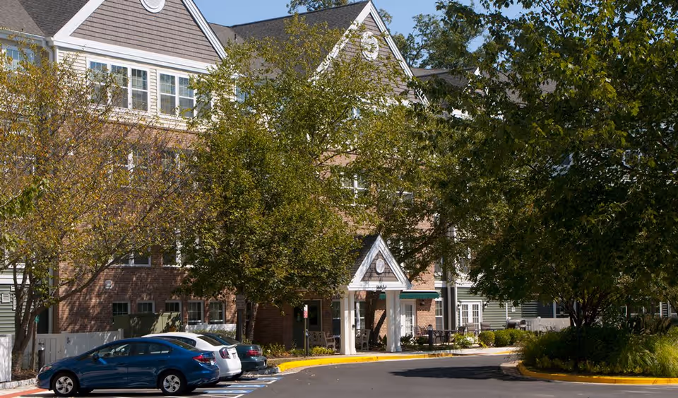 Exterior front entrance of a multi-story brick and siding senior living building with a covered portico, trees, and parked cars.