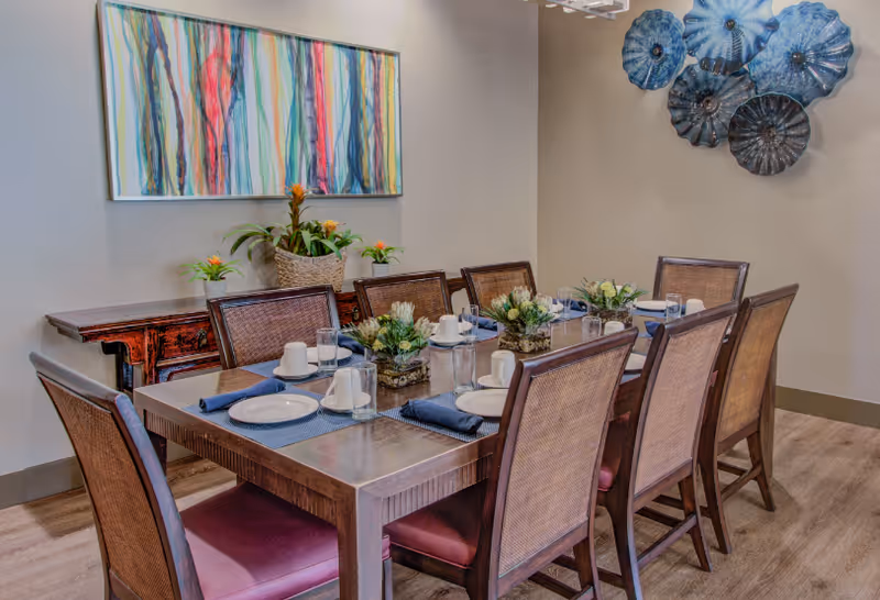 A dining room with a rectangular wooden table set for eight people. The table is arranged with white plates, cups, glasses, and blue napkins. There are three small floral centerpieces on the table. The room has wooden chairs with woven backs and purple cushions. On the wall behind the table, there is a colorful abstract painting and a decorative blue glass wall art installation. A wooden side table with potted plants is also visible against the wall.