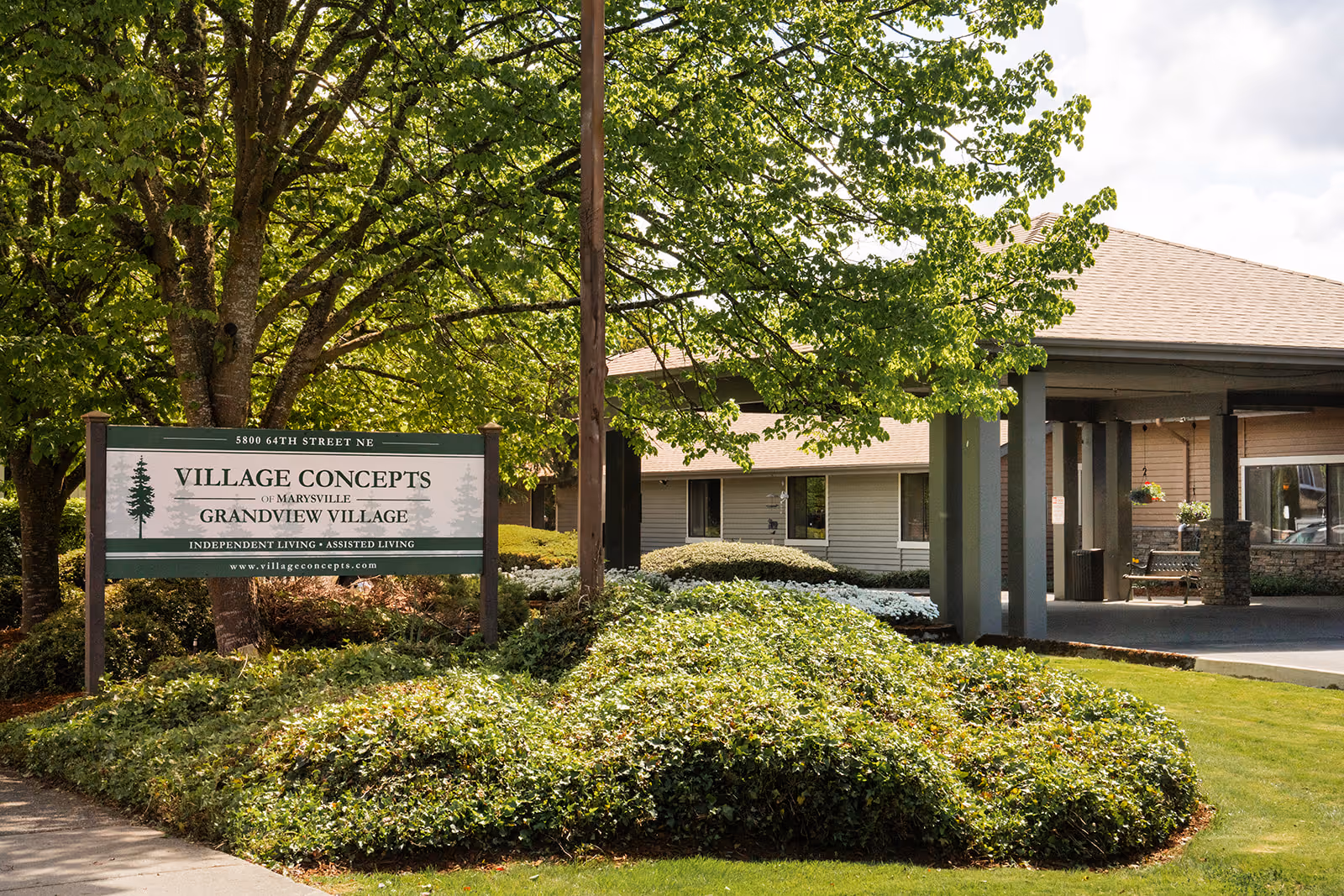 Exterior view of Village Concepts of Marysville - Grandview Village showing the facility's sign surrounded by greenery and trees, with part of the building and covered entrance visible in the background under a partly cloudy sky.