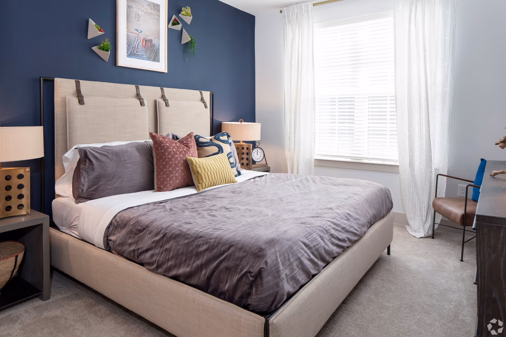 A modern bedroom with a large beige upholstered bed featuring multiple decorative pillows in gray, pink, yellow, and blue. The wall behind the bed is painted dark blue and decorated with a framed bicycle picture and small triangular wall planters with green plants. There are two bedside tables with lamps and a clock on the right side. A window with white blinds and patterned white curtains lets in natural light. A brown chair with metal arms is placed near a dark wooden dresser on the right side of the room.