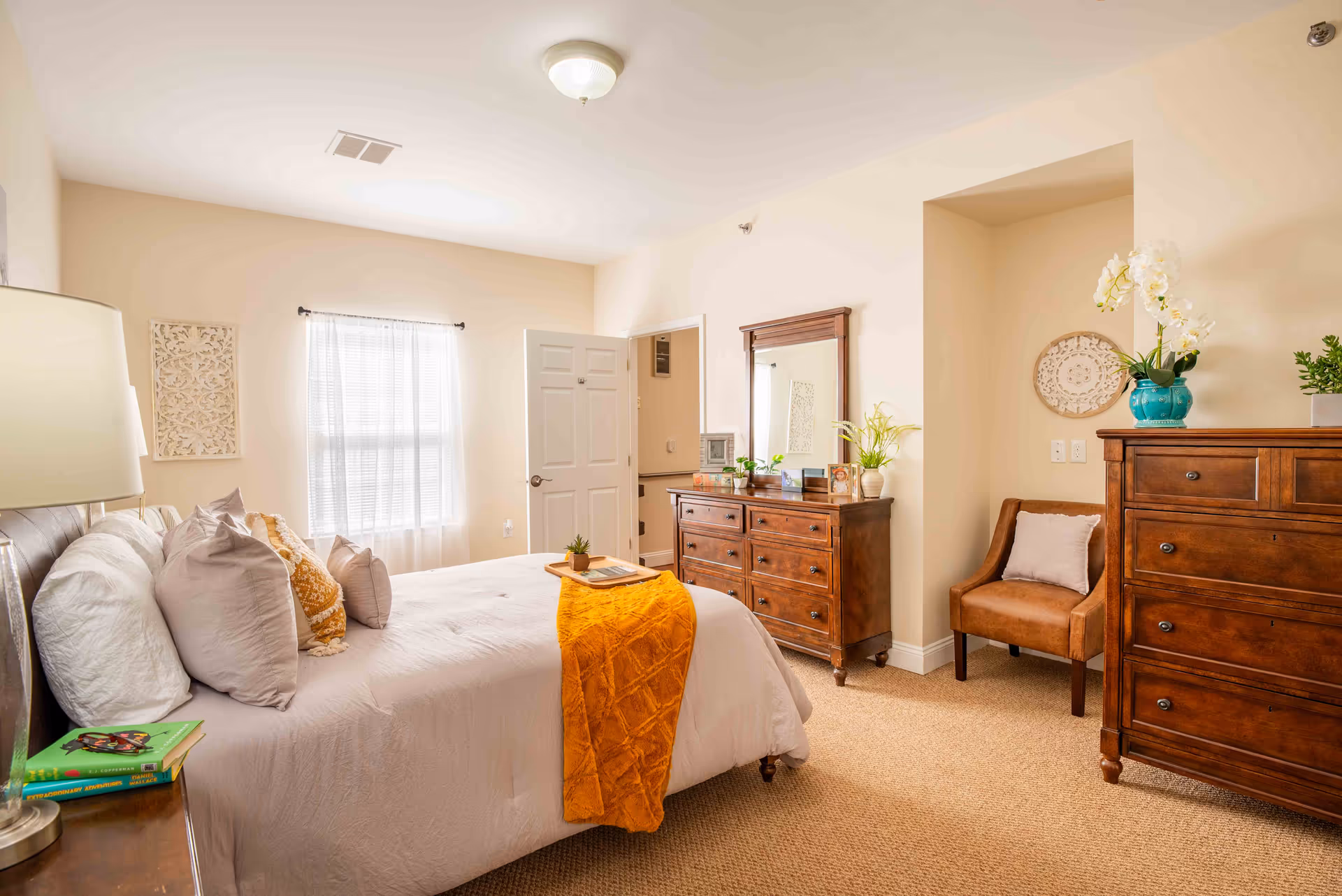 A bright and cozy bedroom with a large bed adorned with multiple pillows and a mustard yellow throw blanket. The room features wooden furniture including a dresser with a mirror, a chest of drawers, and a small upholstered chair in a recessed wall area. There is a window with sheer white curtains allowing natural light to fill the room, and decorative plants and wall art add a touch of greenery and style.