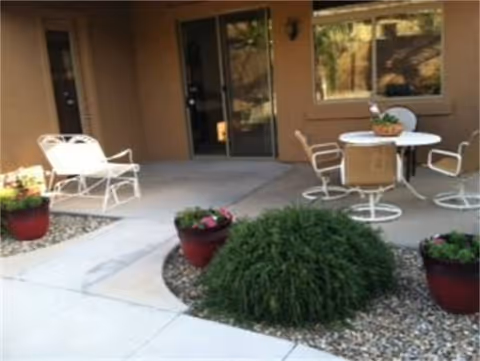 Outdoor patio with white chairs, a round table, potted plants, and a beige building entrance.