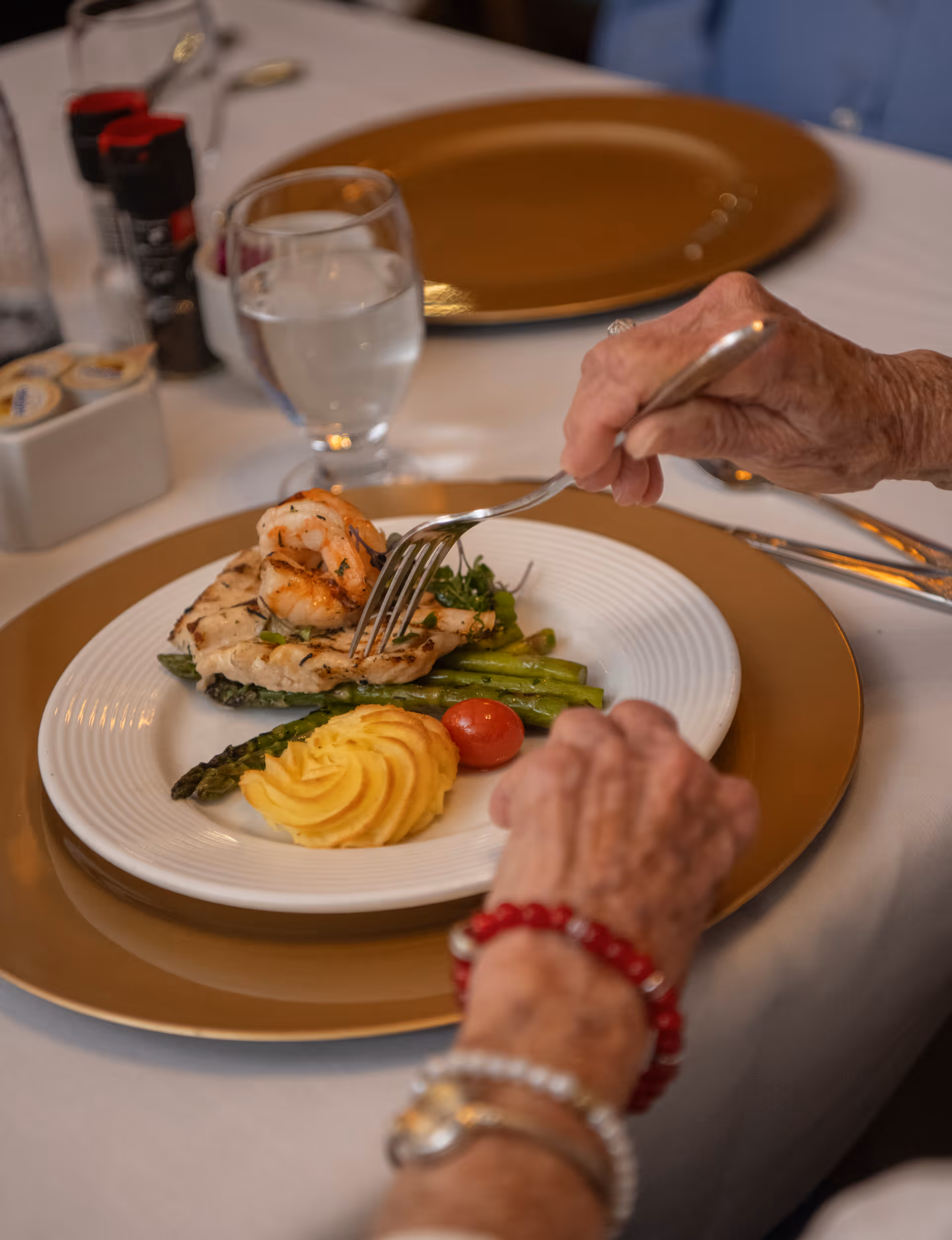 An elderly person wearing bracelets is using a fork to eat a plated meal consisting of grilled shrimp, chicken, asparagus, a cherry tomato, and a serving of mashed potatoes, set on a white tablecloth with a gold charger plate underneath.