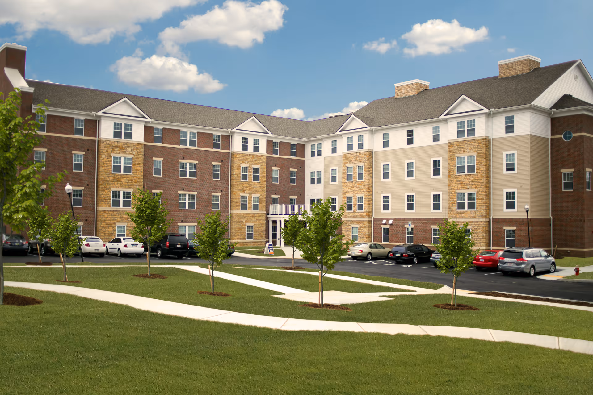 Exterior view of a multi-story senior living facility building with a mix of brick and siding facade, surrounded by a well-maintained lawn with young trees and a paved parking area with several parked cars under a partly cloudy sky.