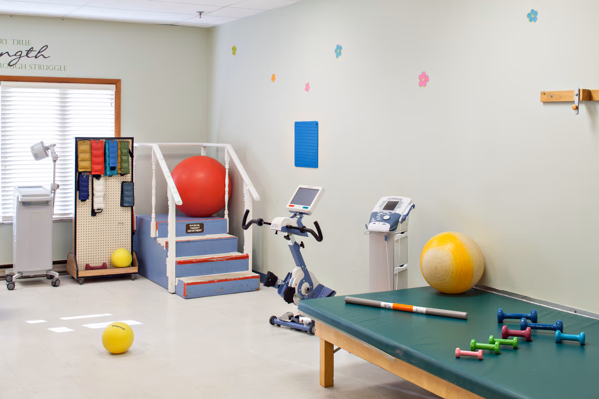 A physical therapy room with exercise equipment including a stationary bike, therapy steps with a large red exercise ball, a yellow exercise ball on the floor, a green therapy table with small colorful dumbbells and a therapy bar, and a pegboard with various exercise bands. The walls are light-colored with small flower decorations.