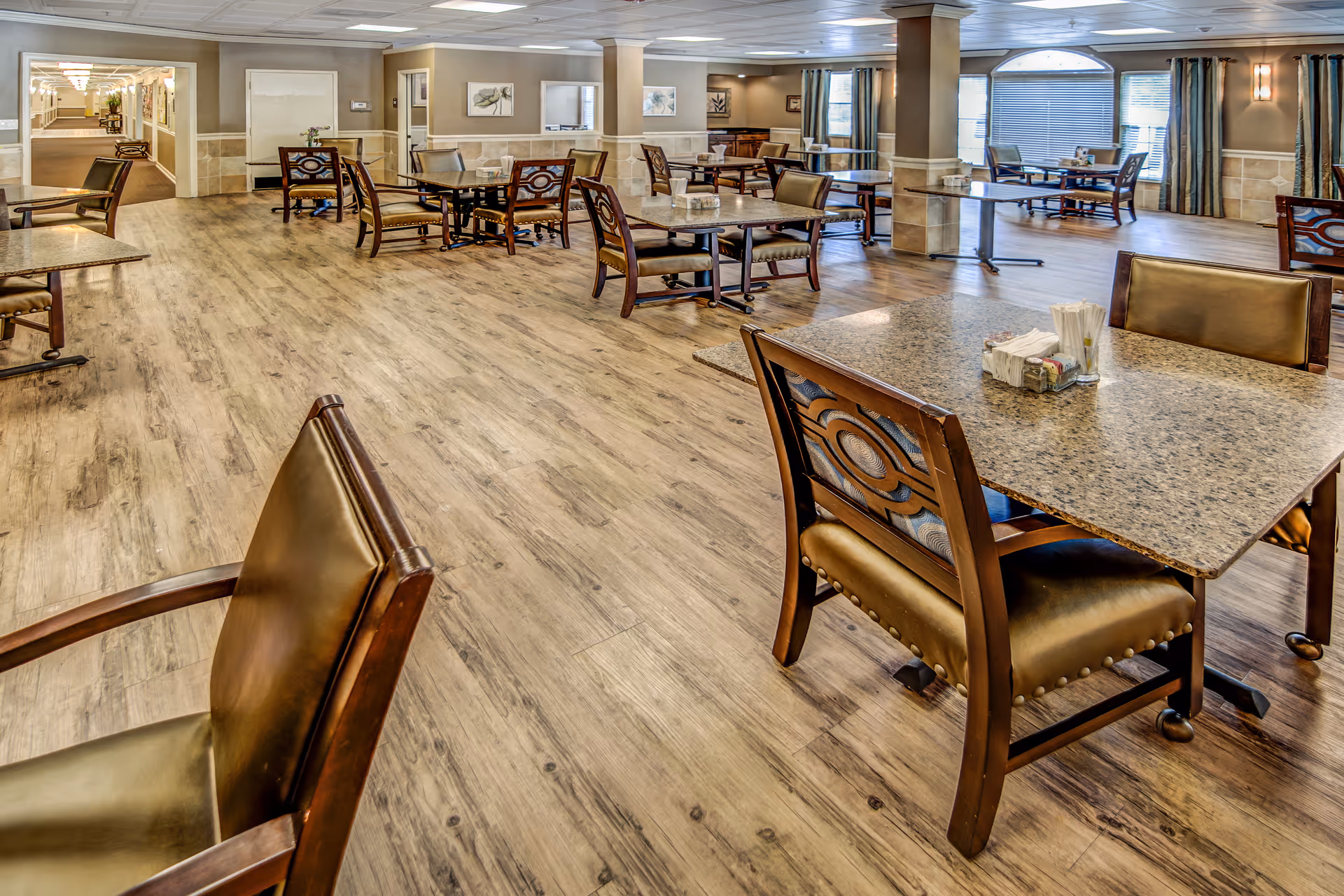 Spacious dining area with multiple tables and chairs arranged on a wood-patterned floor. The room has beige walls with decorative panels, framed artwork, and large windows with curtains allowing natural light. The ceiling has recessed lighting and there is a hallway visible in the background.