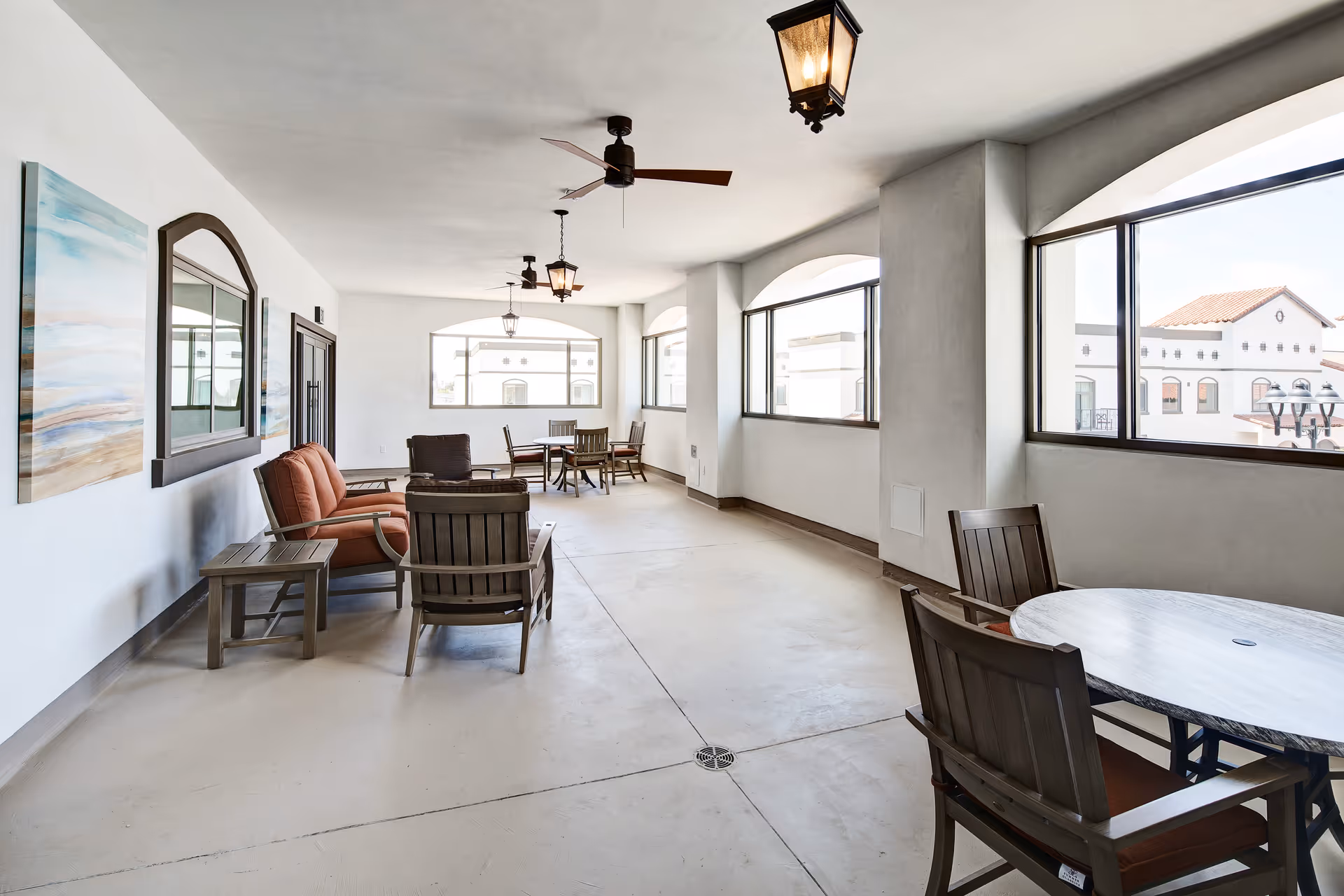 A bright indoor lounge area with large windows showing a view of white buildings with red-tiled roofs. The room features several wooden chairs and tables, a cushioned sofa, ceiling fans, and hanging lantern-style lights. The walls are white with a large mirror and artwork.