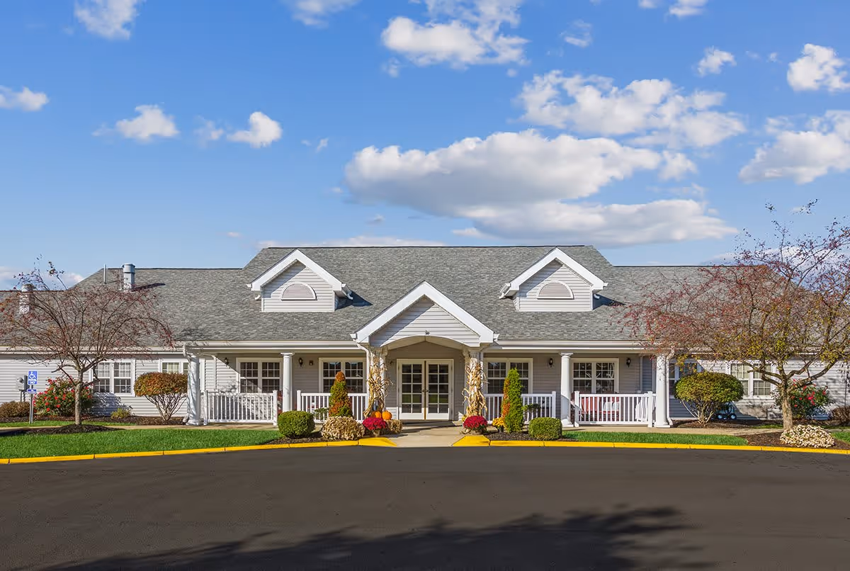 Front exterior view of The Addison of Garden Way Place, a single-story building with a gray roof, white siding, and a covered entrance with columns. The entrance is decorated with fall-themed plants and shrubs, and there are small trees and bushes on either side of the building. The sky is blue with scattered clouds.
