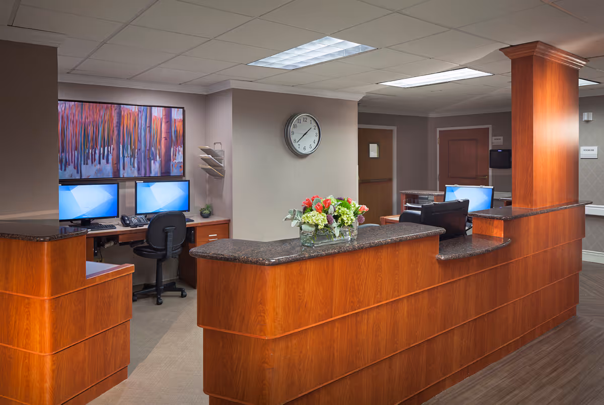 Reception area with a wooden desk featuring a granite countertop, a vase with flowers, two computer workstations with chairs, a wall clock showing 3:10, and a colorful abstract painting on the wall.