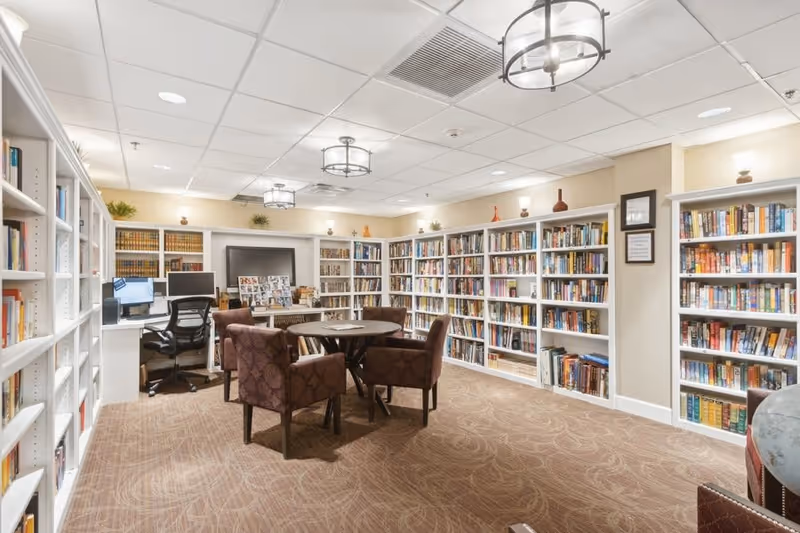A well-lit library room in a senior living facility with white bookshelves filled with books lining the walls. There are four upholstered chairs around a circular table in the center of the room. A desk with a computer and office chair is positioned against one wall. The ceiling has recessed lighting and decorative hanging light fixtures.