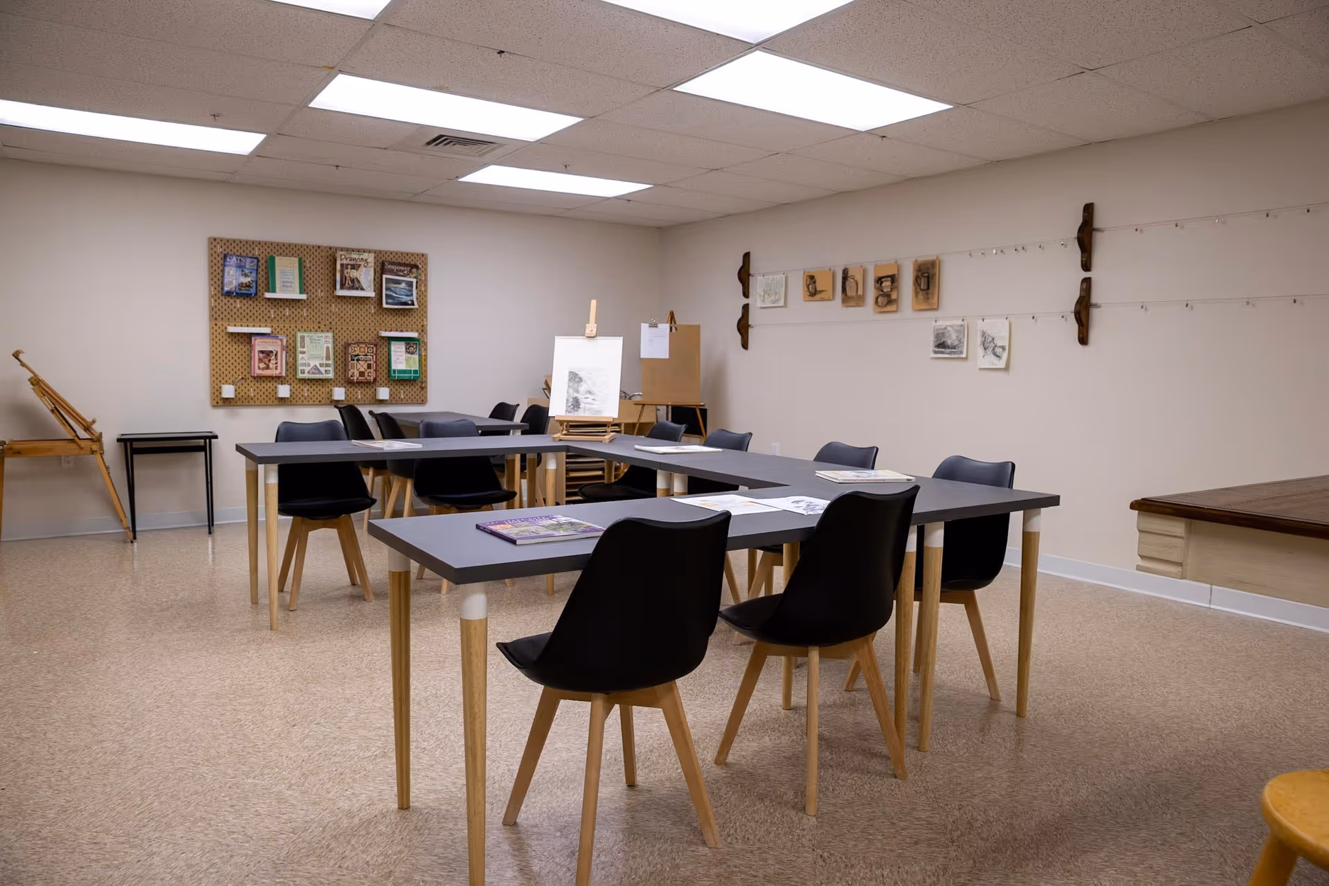A well-lit art room with several black chairs around gray tables arranged in a U-shape. The walls display various sketches and drawings, with an easel holding a drawing pad. The floor is covered with light-colored linoleum, and the ceiling has fluorescent lights.
