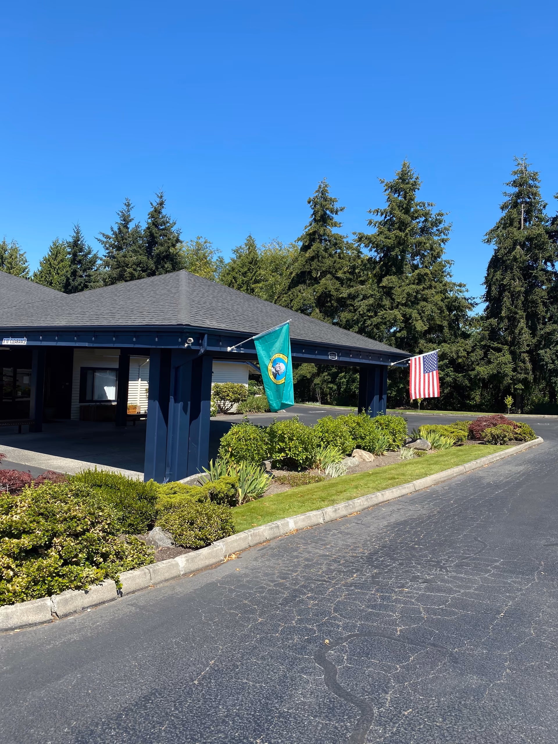 Exterior view of Logan Creek Retirement Community entrance with a covered driveway, landscaped bushes, and two flags - one green state flag and one American flag, under a clear blue sky with tall evergreen trees in the background.