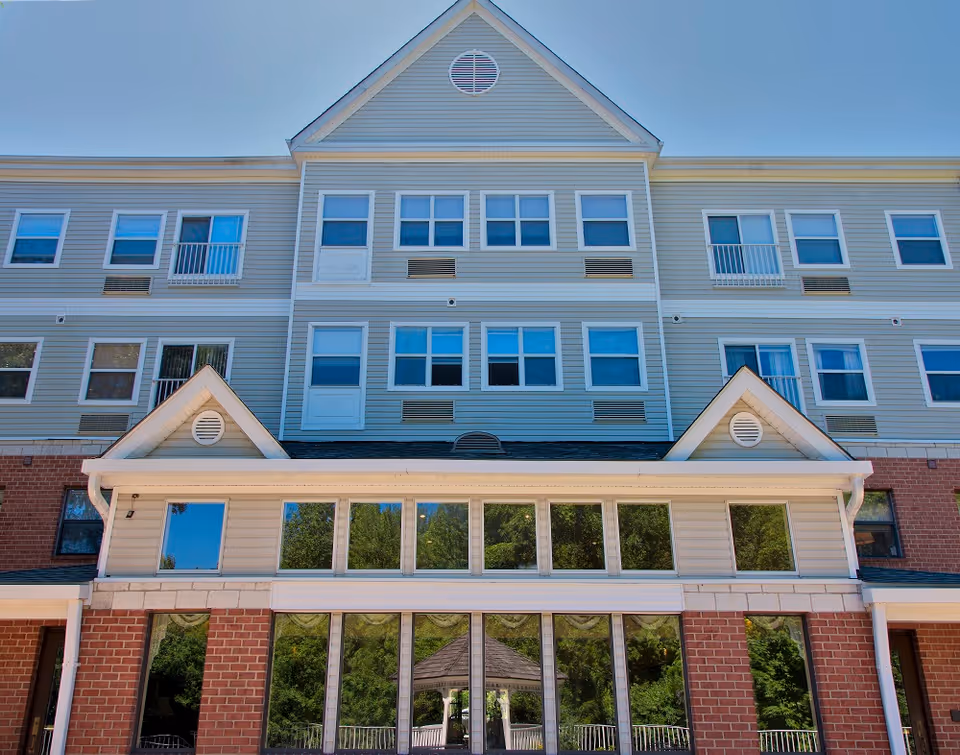 Exterior view of a multi-story senior living facility building with beige siding and red brick on the lower level. The building has multiple windows and a peaked roof section. Reflections of trees and a gazebo are visible in the large windows on the ground floor.