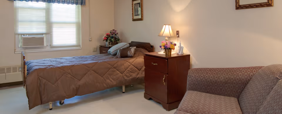 Small furnished bedroom with a single bed, nightstand with a lamp and flowers, a window air-conditioner, and a sofa.