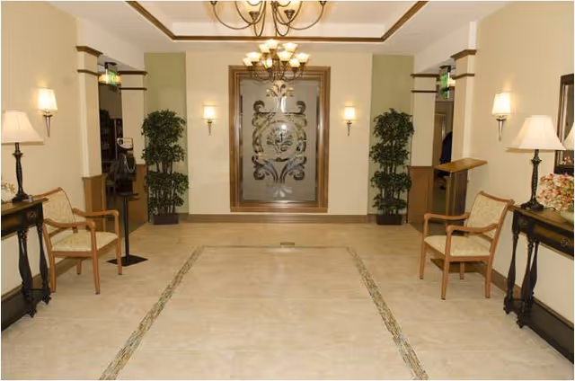 Well-lit lobby with a decorative frosted glass door, chairs, side tables, potted plants, and chandeliers.
