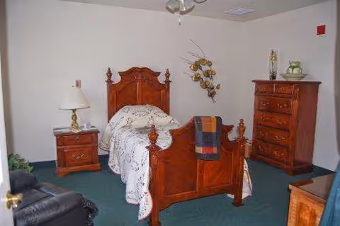 A bedroom with a wooden single bed featuring a carved headboard and footboard, a floral bedspread, a plaid blanket draped over the footboard, a wooden nightstand with a lamp, a wooden dresser with decorative items on top, a black recliner chair, and a wall decoration with metal leaves.