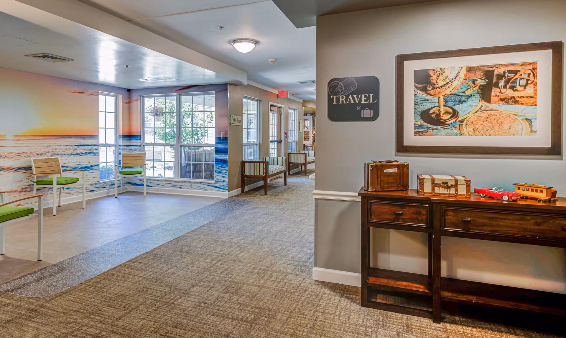 Interior hallway of Arden Courts - ProMedica Memory Care Community with a travel-themed decor. The right side features a wooden console table with decorative vintage suitcases and toy vehicles. Above the table is a framed picture of a globe and maps. A sign on the wall reads 'TRAVEL' with travel-related icons. The left side has a mural of a sunset over the ocean with chairs lined up against the wall. The hallway has carpeted flooring and leads to a door with windows letting in natural light.