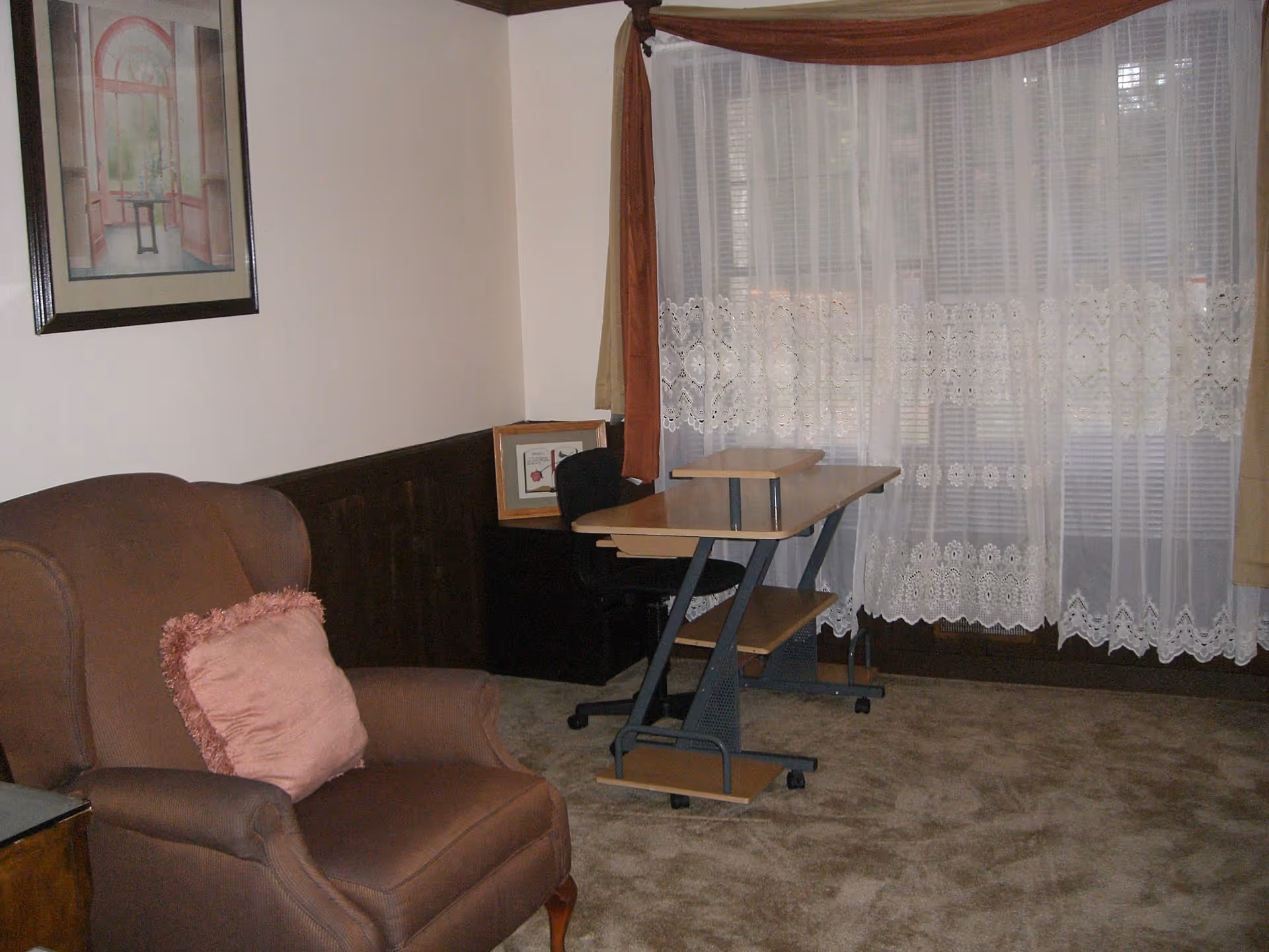 A cozy room with a brown upholstered armchair featuring a pink decorative pillow, a small wooden desk with a black office chair, and a large window covered with sheer white lace curtains and brown drapes. A framed picture hangs on the wall above the armchair.