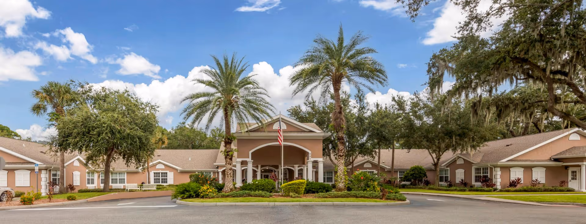 Single-story senior living facility entrance framed by palm trees, a landscaped circular driveway, and an American flag under a blue sky.