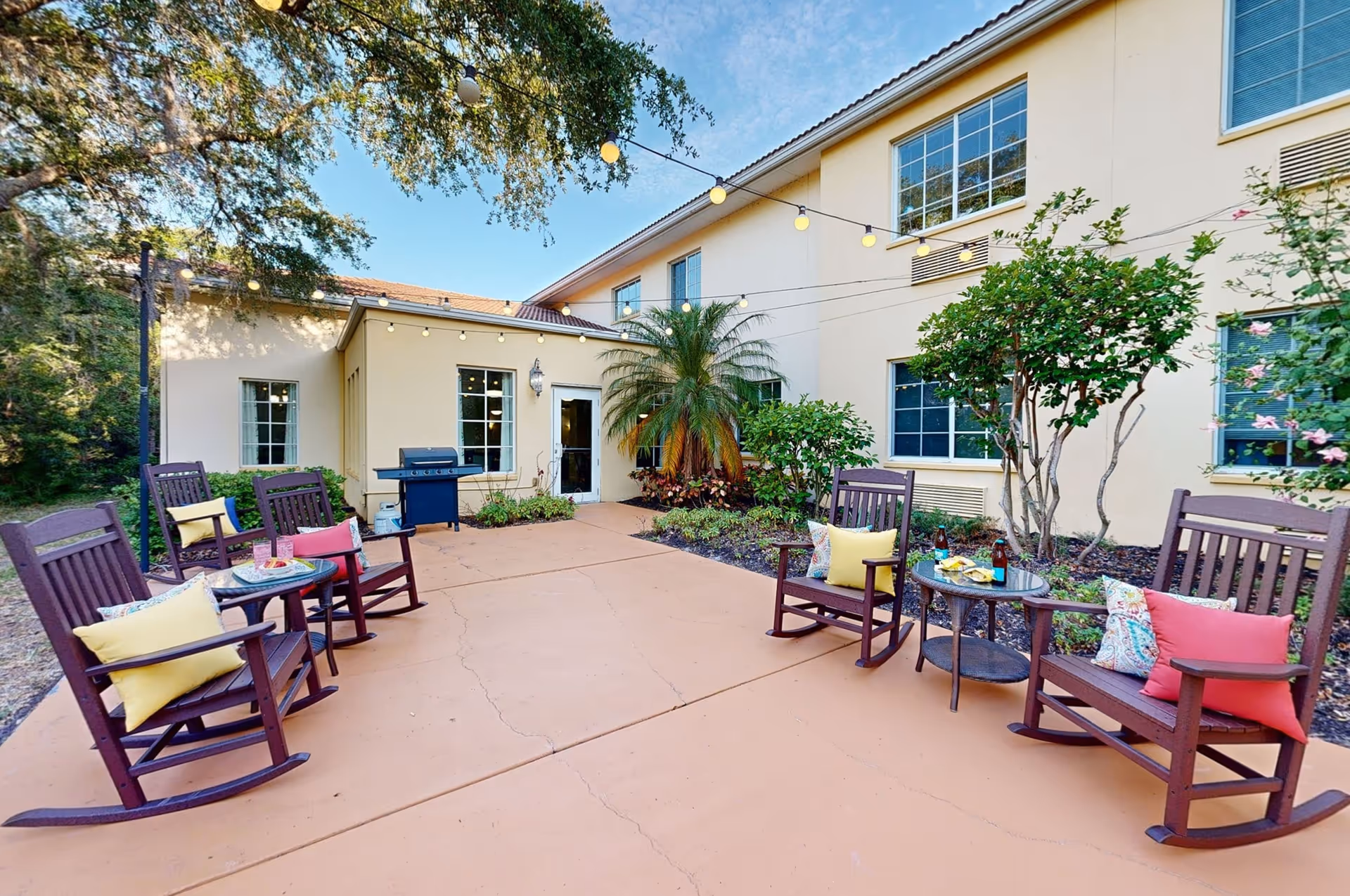 Outdoor patio area at The Windsor of Lakewood Ranch featuring several wooden rocking chairs with colorful cushions arranged around small round tables. String lights are hung overhead, and there are plants and trees along the building's exterior walls.