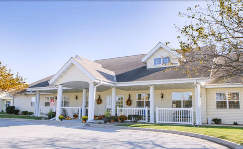 Single-story light-yellow senior living building with a covered porch, white columns, and potted plants at the entrance under a clear sky.
