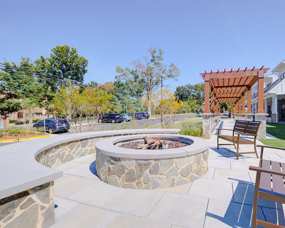 Sunny outdoor courtyard featuring a circular stone fire pit, seating benches, and a wooden pergola.