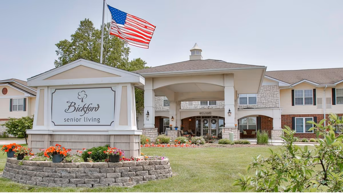 Front entrance of Bickford senior living with a large sign, landscaped flower beds, and an American flag flying above.