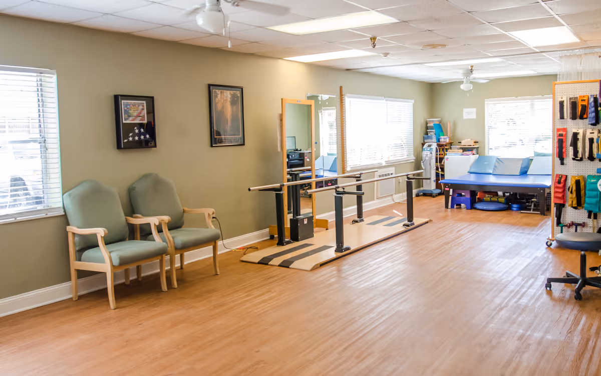 A bright rehabilitation therapy room with parallel walking bars, seating, and exercise tables.
