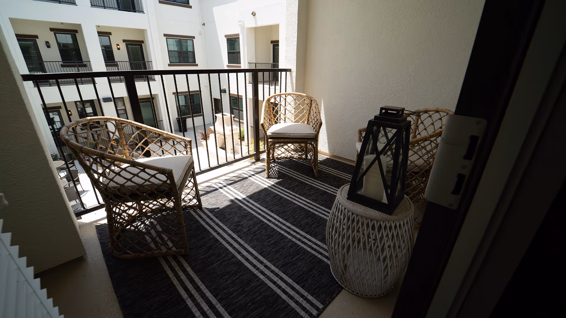 A small balcony with three wicker chairs with cushions arranged around a wicker side table holding a black lantern with a candle inside. The balcony has a black and white striped rug and overlooks the courtyard of a multi-story building with white walls and multiple windows.