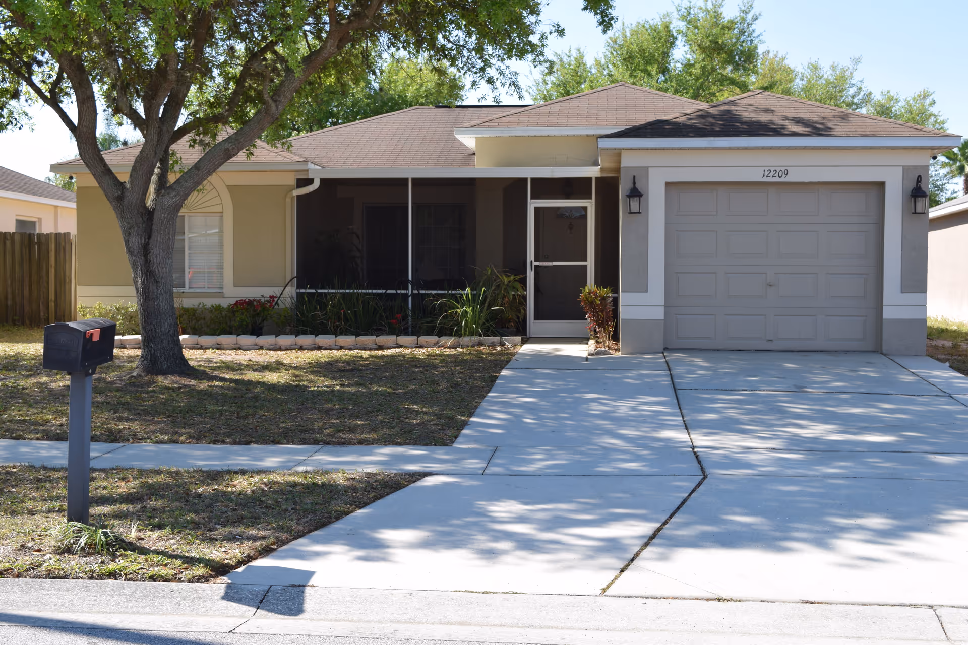Front exterior view of a single-story residential house with a brown shingle roof, beige walls, a garage door, a screened front porch, a tree in the front yard, a mailbox near the sidewalk, and a concrete driveway.