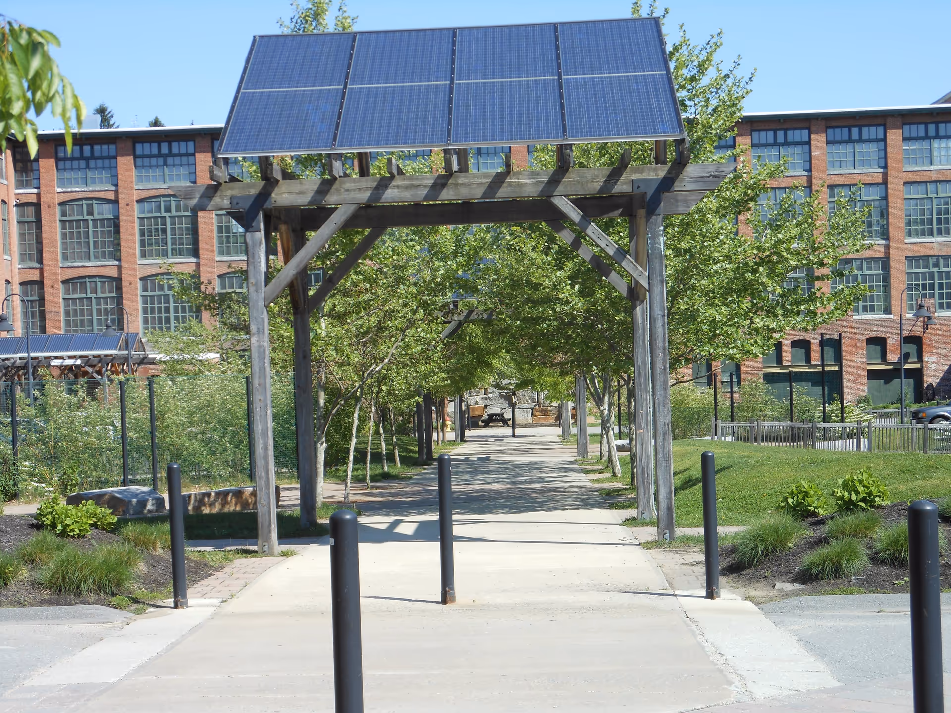 A tree-lined paved walkway passes under a wooden pergola topped with solar panels toward a large brick building.