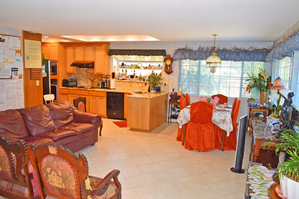 A bright and cozy open living space featuring a kitchen with wooden cabinets and granite countertops, a dining area with a table covered in a floral tablecloth and red chair covers, and a seating area with a brown leather couch and ornate wooden chairs. Large windows with blue valances let in natural light, and various plants and decorative items are placed throughout the room.
