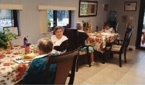 A dining area in a senior living facility with several elderly individuals seated around tables covered with floral tablecloths. The room has large windows with blinds, framed pictures on the walls, and a tiled floor.