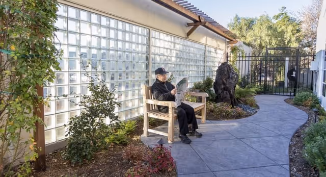 An elderly man wearing a black jacket and cap is sitting on a wooden bench reading a newspaper in a garden courtyard area. The courtyard has a curved concrete pathway, plants, and bushes along the sides, and a glass block wall on one side with a metal gate at the end of the path.
