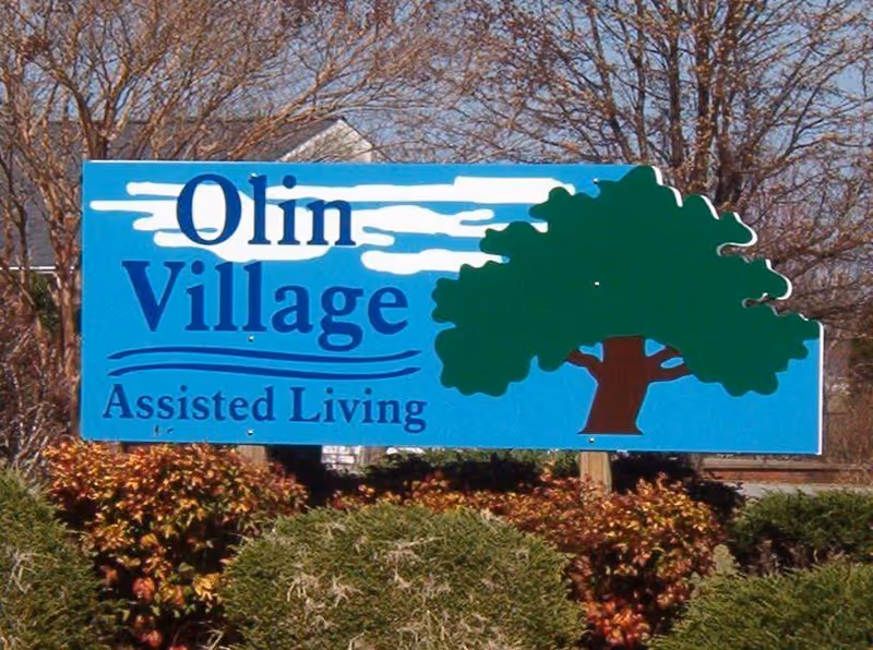 Blue roadside sign that reads "Olin Village Assisted Living" with a large green tree graphic and shrubs in front.