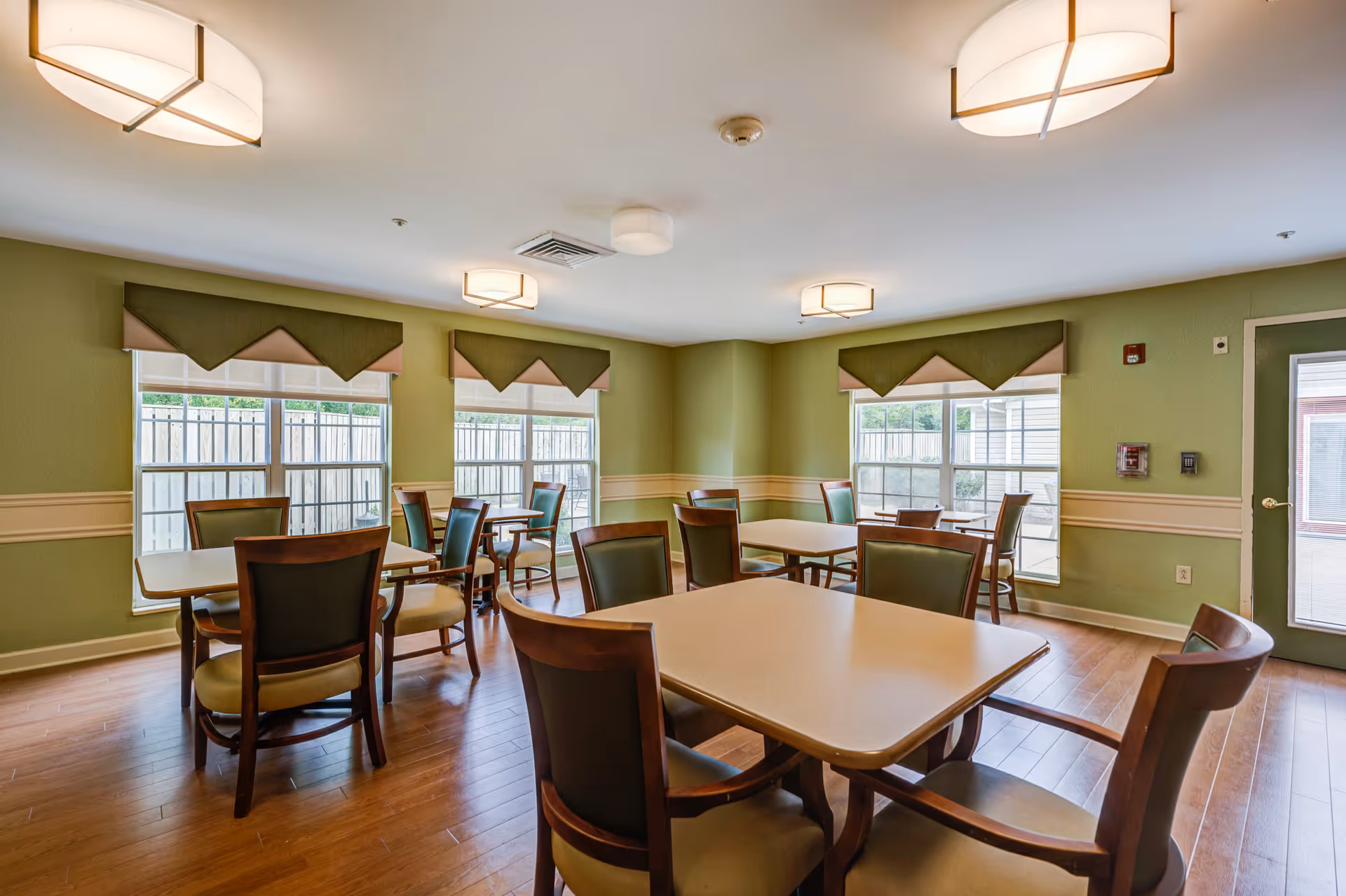 A bright dining room with several square tables and wooden chairs with green cushions. The walls are painted light green with a beige chair rail, and there are large windows with green and beige valances letting in natural light. The floor is wood, and modern ceiling lights illuminate the room.