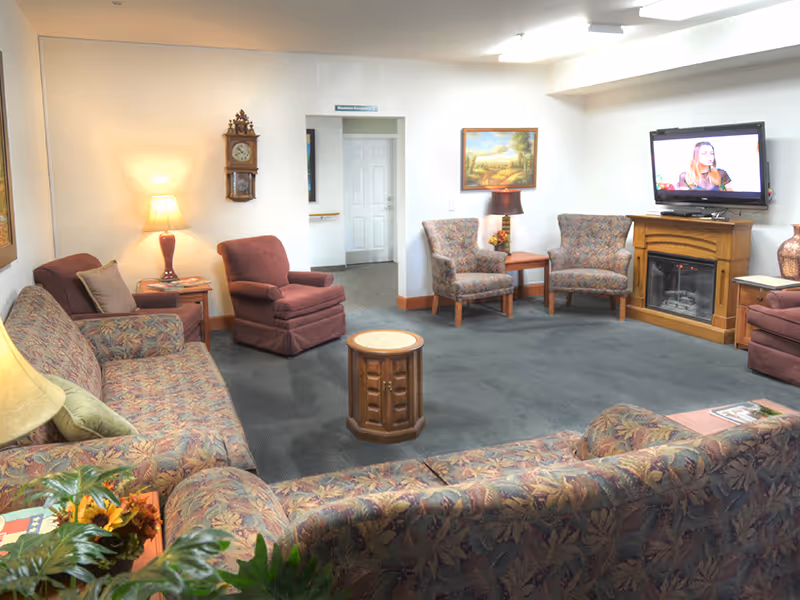 A cozy senior living community lounge with patterned sofas and armchairs arranged around a small wooden table. There is a wall-mounted TV above an electric fireplace, a grandfather clock on the wall, and a couple of table lamps providing warm lighting. A painting hangs on the wall, and a doorway leads to another room.