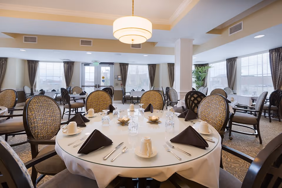 A dining room in a senior living facility with round tables set for a meal. Each table has a white tablecloth, neatly folded dark napkins, cups, glasses, and silverware. The room is well-lit with large windows covered by curtains, and there is a modern ceiling light fixture above the central table.