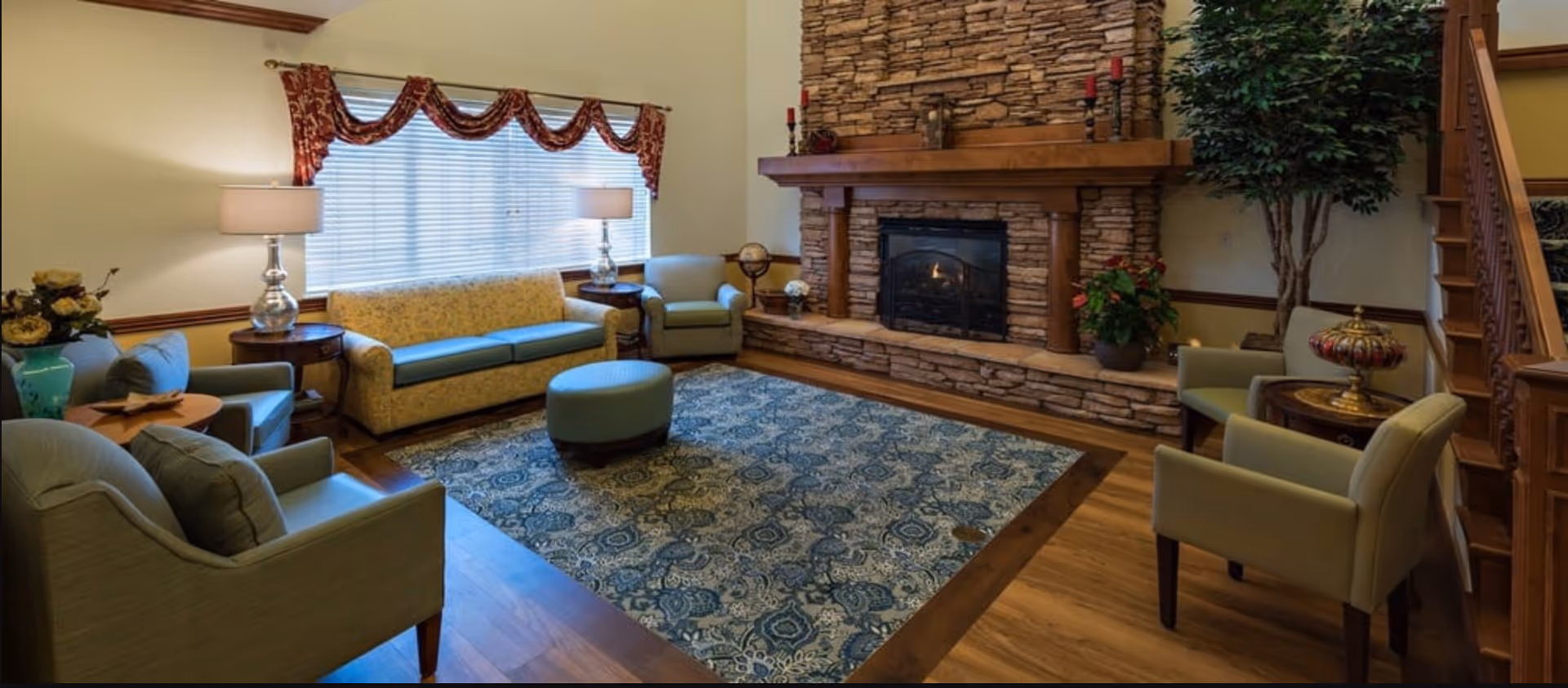 A cozy living room with a stone fireplace, a patterned area rug, a floral sofa, several armchairs, two table lamps, decorative plants, and a wooden staircase.