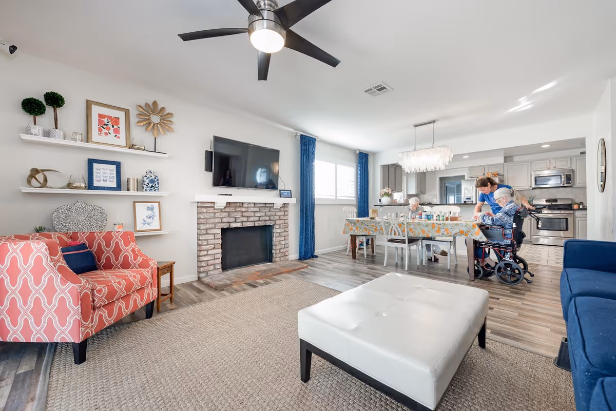 A bright and spacious living and dining area in a senior care home. The room features a red patterned armchair, a white ottoman, a blue sofa, and a brick fireplace with a mounted flat-screen TV above it. Shelves with decorative items are on the wall to the left. In the dining area, an elderly person in a wheelchair is being assisted by a caregiver at a table covered with a floral tablecloth. The kitchen with modern appliances is visible in the background.