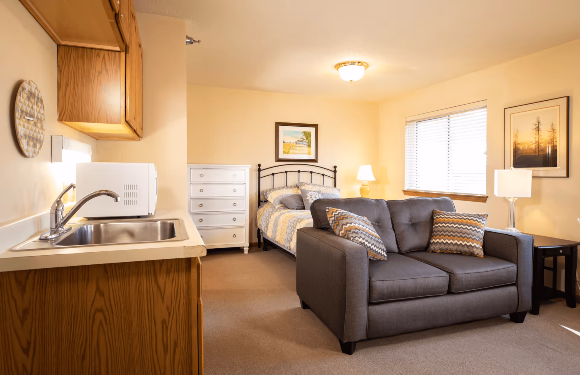A cozy assisted living studio apartment featuring a small kitchenette with a sink and microwave on the left, a gray loveseat with patterned pillows in the center, and a bed with a metal headboard and floral bedding against the back wall. There is a white dresser next to the bed, two table lamps on either side, and framed artwork on the walls. A window with blinds allows natural light into the room.