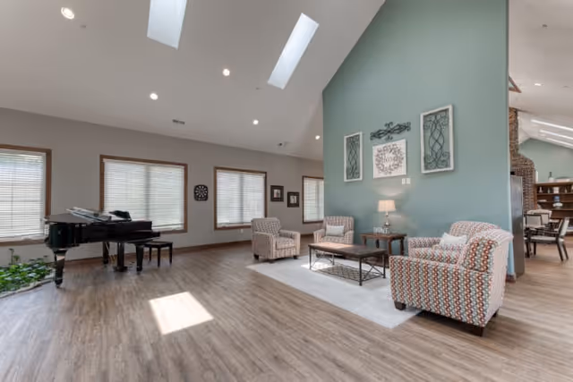 A spacious and well-lit living room area with vaulted ceilings and skylights. The room features three patterned armchairs arranged around a coffee table on a light-colored rug. A side table with a lamp and decorative wall art are visible on a teal accent wall. Large windows with blinds allow natural light to fill the space. A black grand piano is positioned near the windows, and a dining area with chairs and shelves is visible in the background.