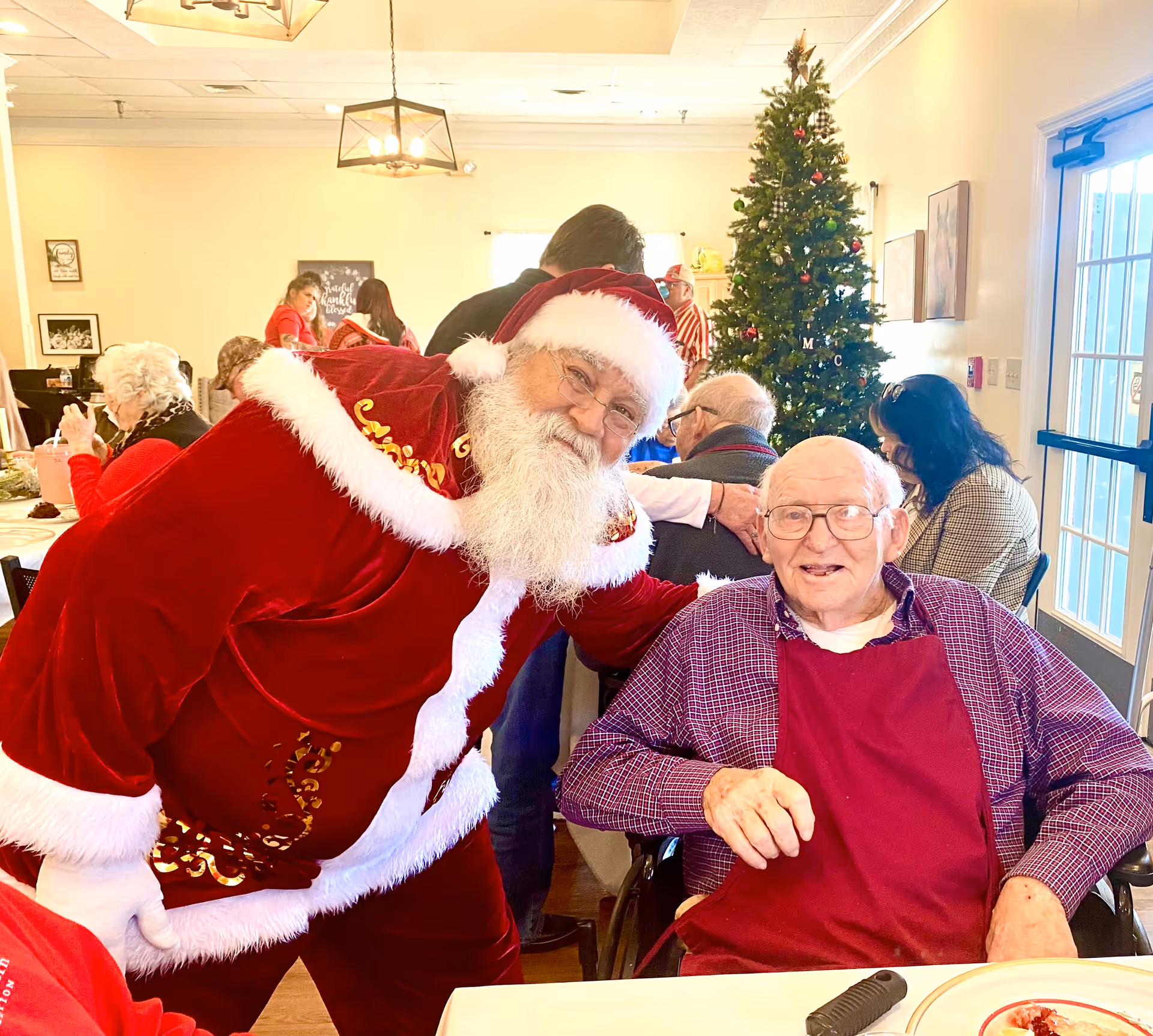 A man dressed as Santa leans in to pose with an elderly resident at a decorated communal dining room with a Christmas tree.