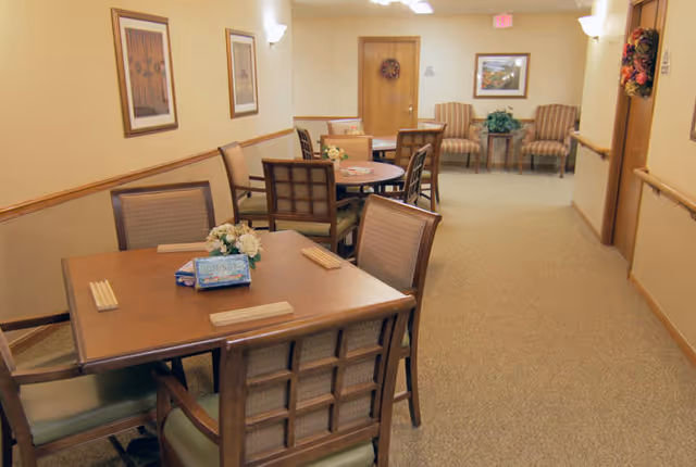 Interior view of a common area in a senior living facility with multiple tables and chairs arranged for seating. The walls are decorated with framed pictures and wreaths, and there are two armchairs with a small table and plant in the background. The space is well-lit with wall-mounted lights.