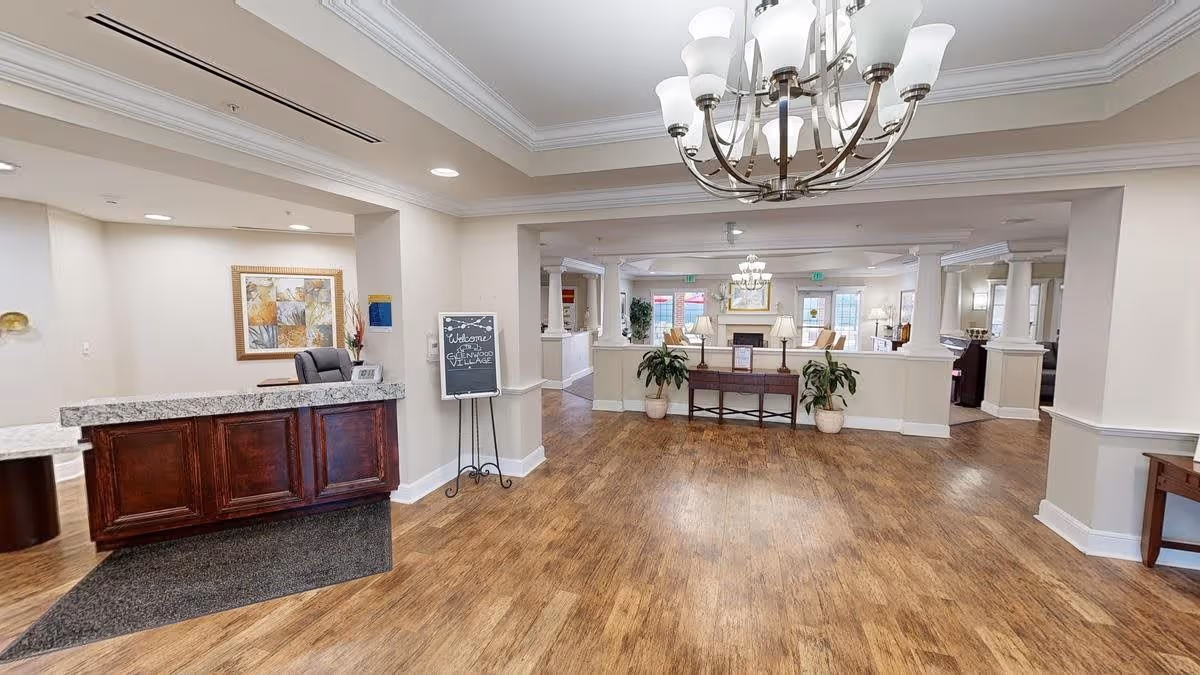 Interior view of a senior living facility lobby with a wooden reception desk on the left, a welcome sign on an easel, hardwood flooring, and a chandelier hanging from the ceiling. In the background, there is a seating area with lamps, plants, and large windows letting in natural light.