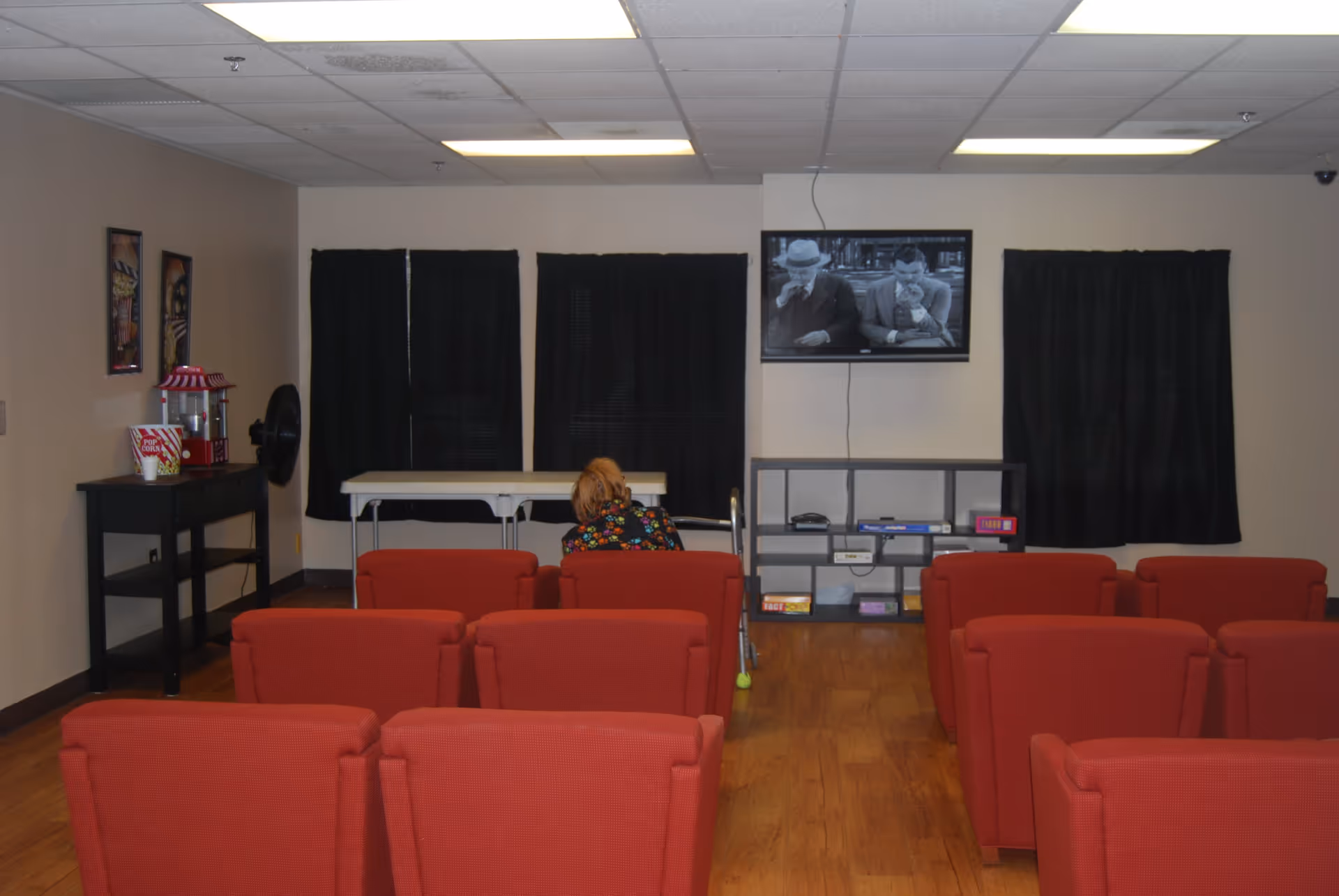 A small movie or TV viewing room with rows of red upholstered chairs facing a wall-mounted flat screen TV showing a black and white film. A person with a walker is seated in the middle row. The room has wood flooring, black curtains covering the windows, and a popcorn machine on a black table to the left.