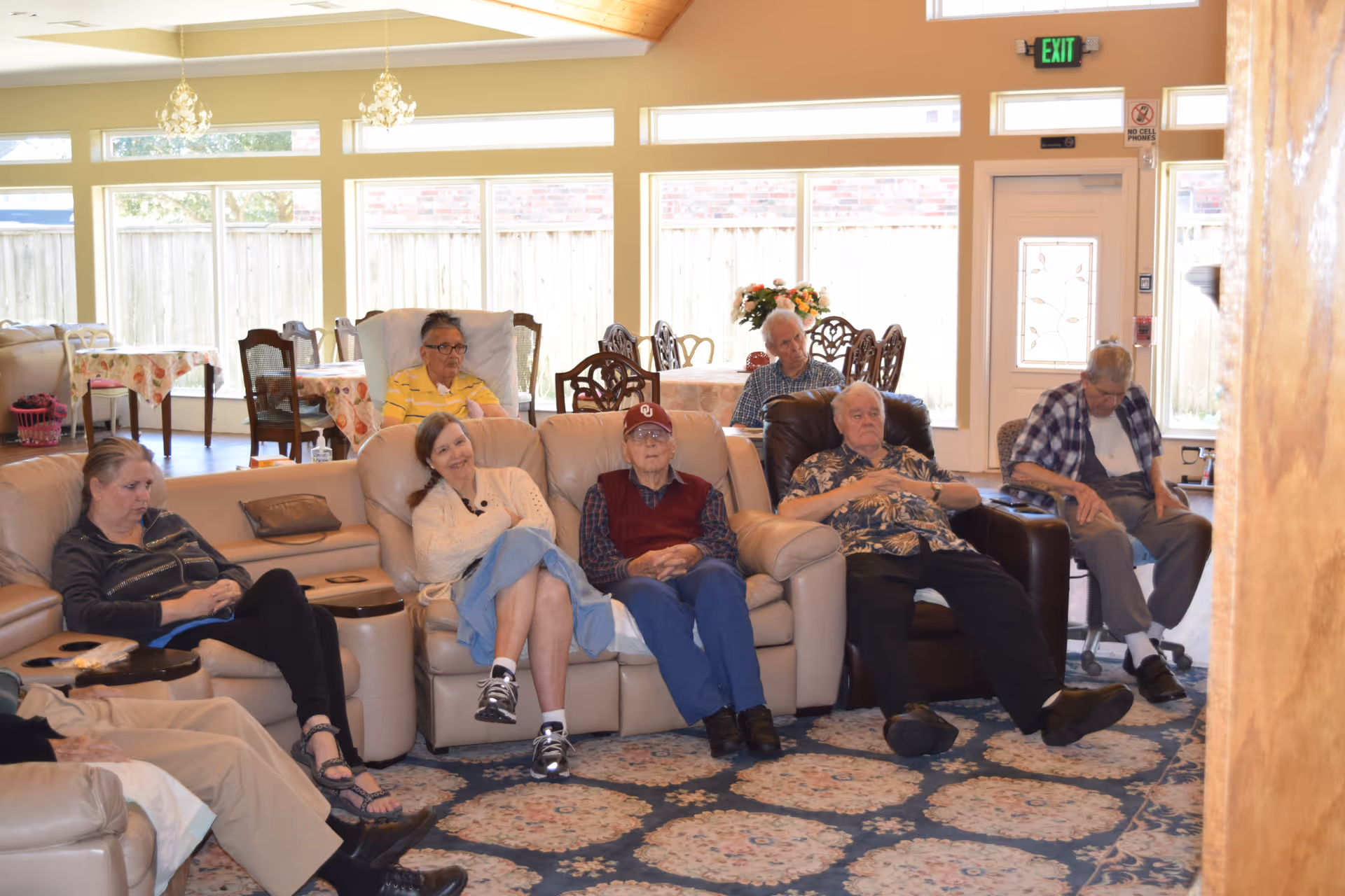 A group of elderly people sitting in a well-lit living room with large windows, comfortable chairs, and a patterned carpet. There are tables with floral tablecloths and a door with an exit sign above it in the background.